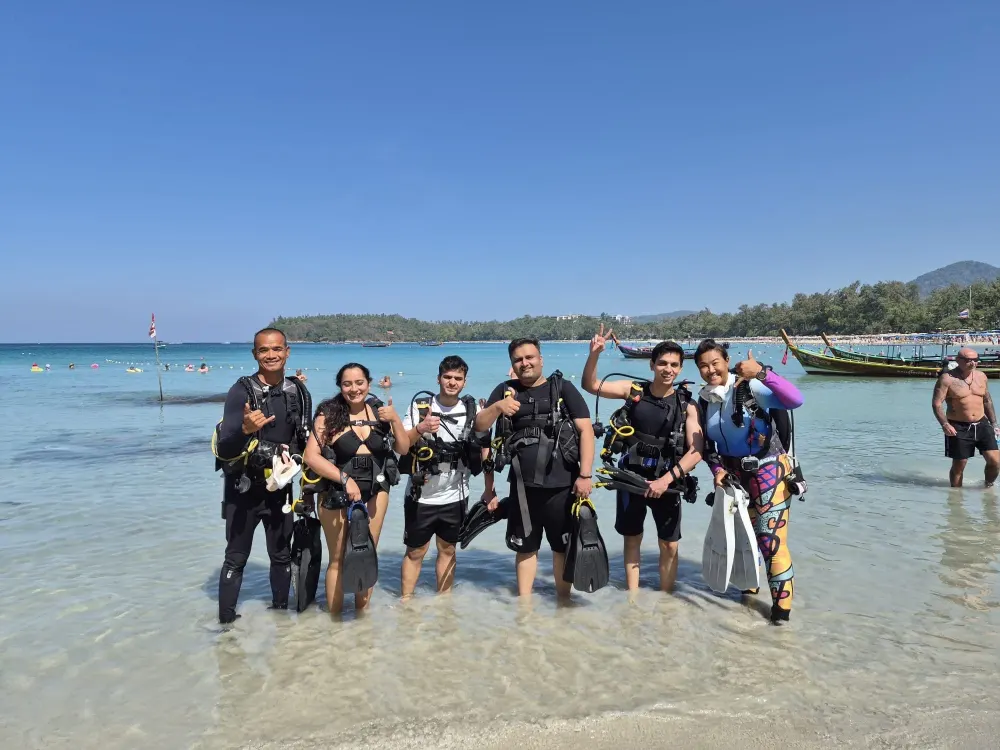 Group of smiling scuba divers in full gear giving thumbs up while standing in the shallows at Kata Beach, Phuket